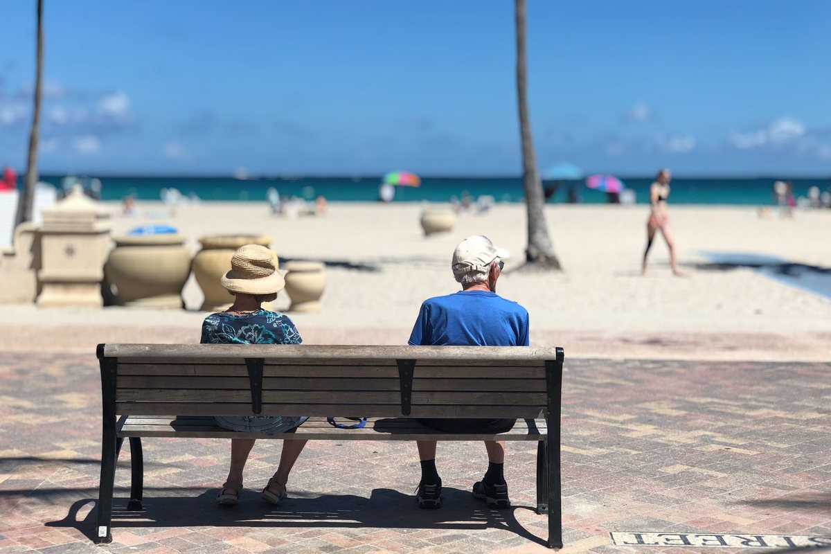 Retired couple sitting together on a wooden bench facing a sunny beach with palm trees