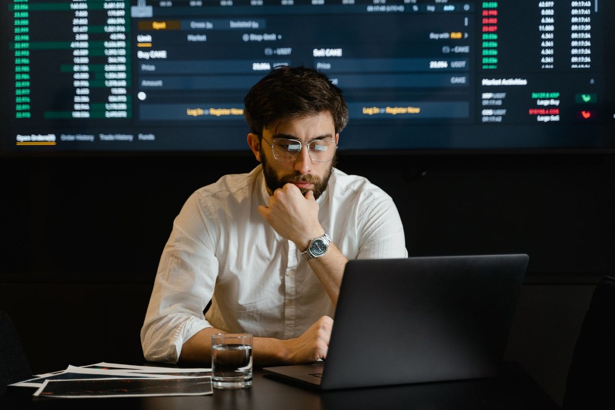 Man focused on a laptop screen showing financial charts while reviewing market data at a desk